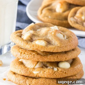 A stack of soft, chewy white chocolate macadamia cookies with golden edges. With a glass of milk and extra plate of cookies in the background.
