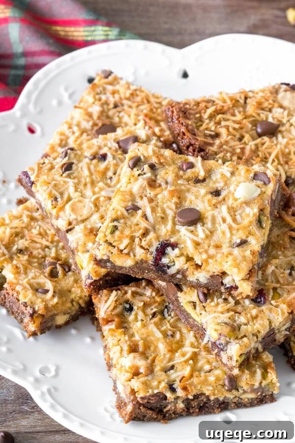 A plate of Christmas magic cookie bars cut into squares and stacked, showcasing the layers of gingersnap crust, cranberries, white chocolate, pistachios, and coconut.