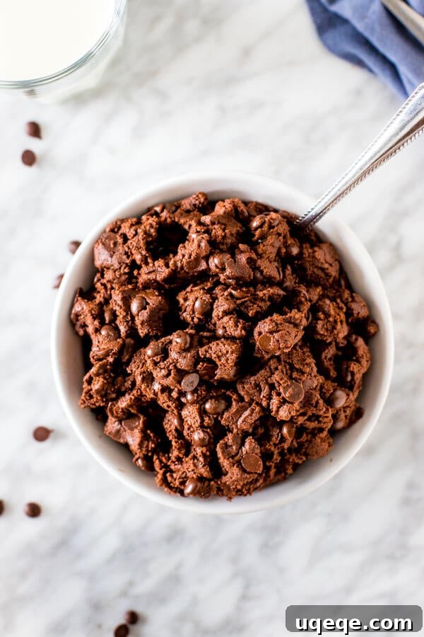 A close-up, overhead shot of rich, dark eggless chocolate cookie dough resting in a rustic ceramic bowl, highlighting its dense, fudgy texture and embedded chocolate chips.