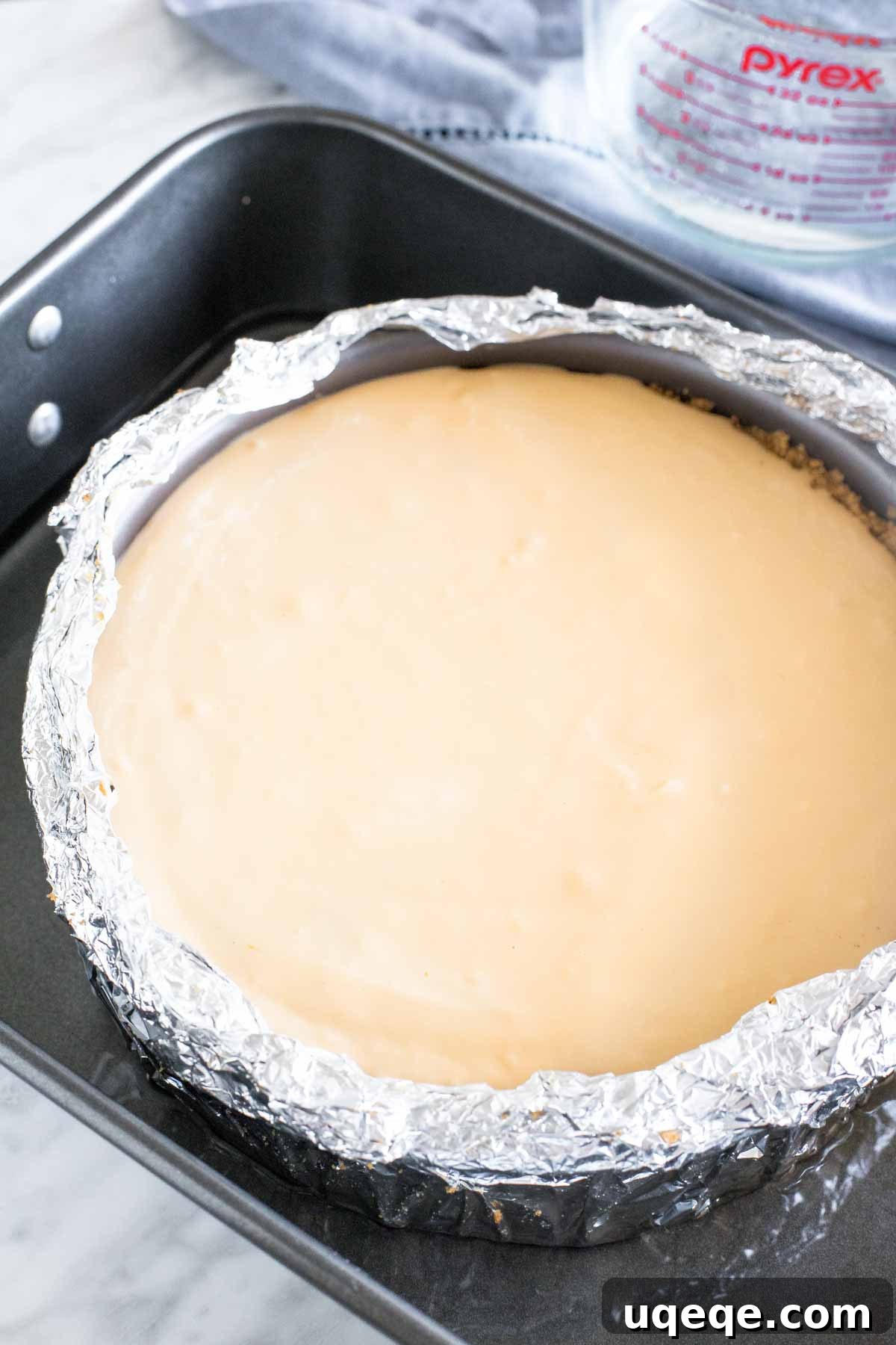 An unbaked cheesecake in a springform pan, wrapped in aluminum foil, positioned inside a larger roasting pan with water.