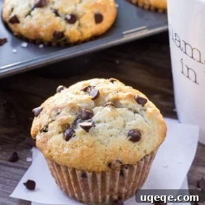 One big, fluffy bakery-style chocolate chip muffin with a cup of coffee and pan full of muffins in the background.