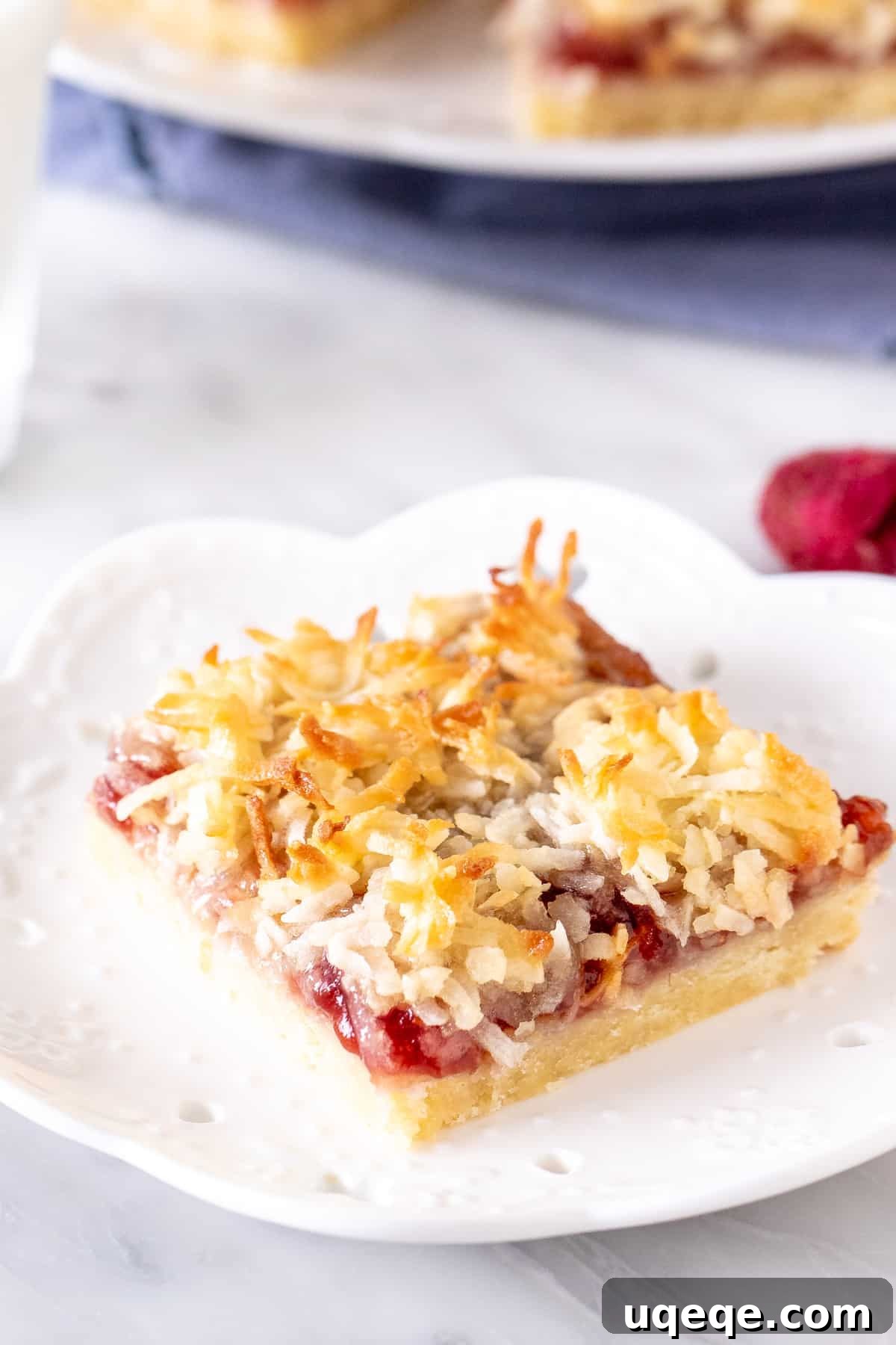 Raspberry Coconut Delights 2 Raspberry coconut slice on a plate, showcasing the distinct layers of shortbread, jam, and coconut. The top is golden brown.