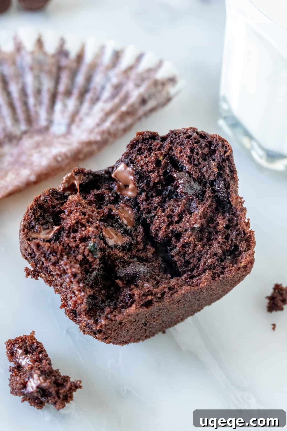 A close-up shot of a chocolate zucchini muffin cut in half, revealing its moist crumb and scattered chocolate chips, with a glass of milk in the background.