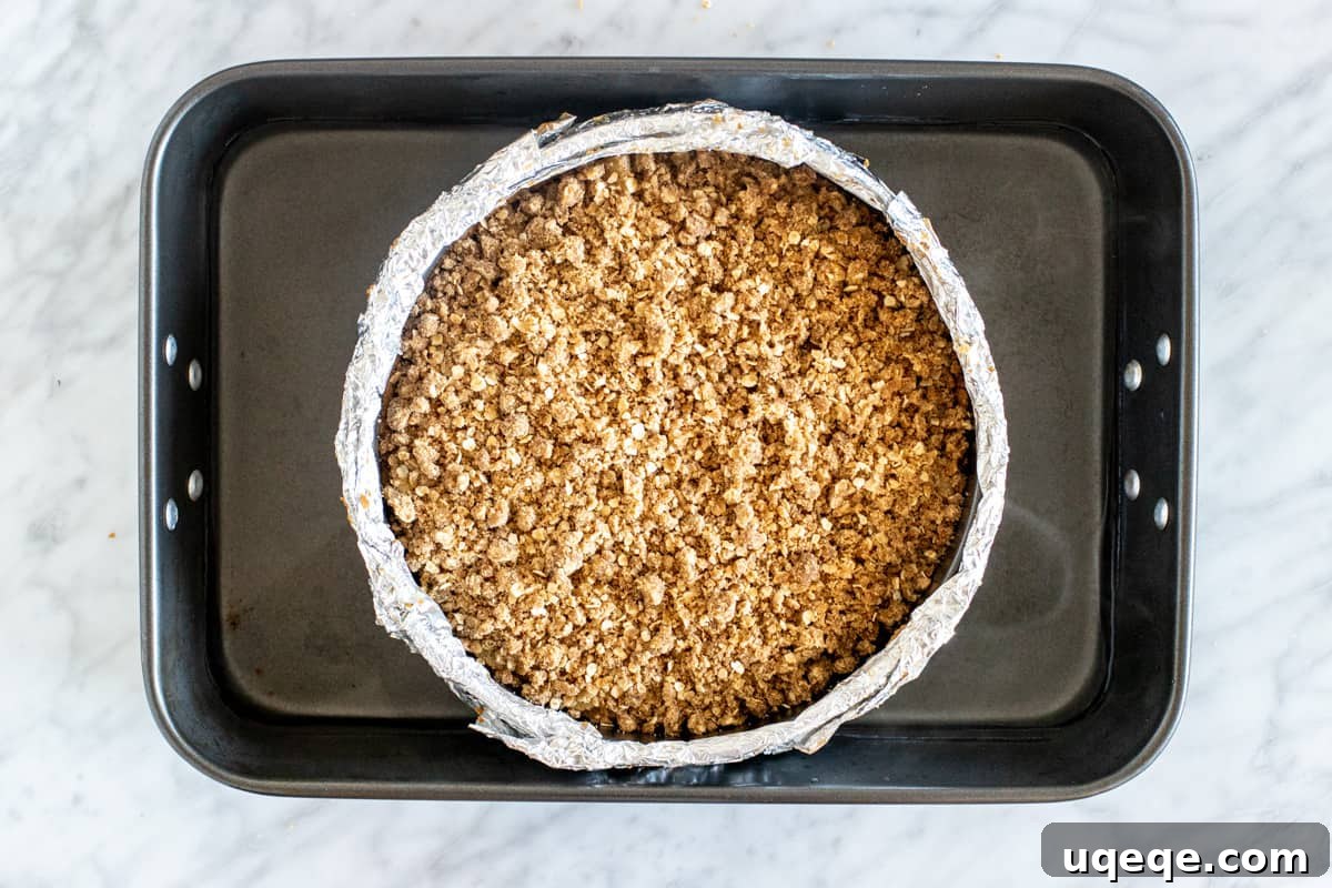 The unbaked Apple Crisp Cheesecake nestled in its foil-wrapped springform pan, placed inside a larger roasting pan filled with boiling water, demonstrating the water bath technique.