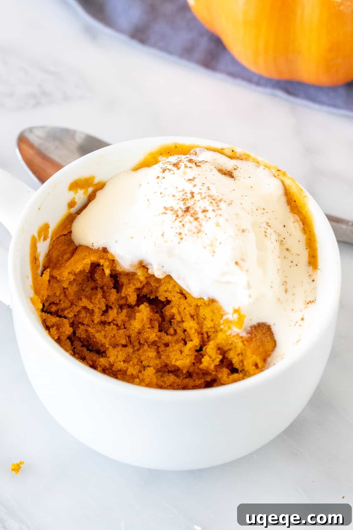 A pumpkin cake in a white mug, with a small bite taken out and topped with luscious melted whipped cream, showcasing its moist texture