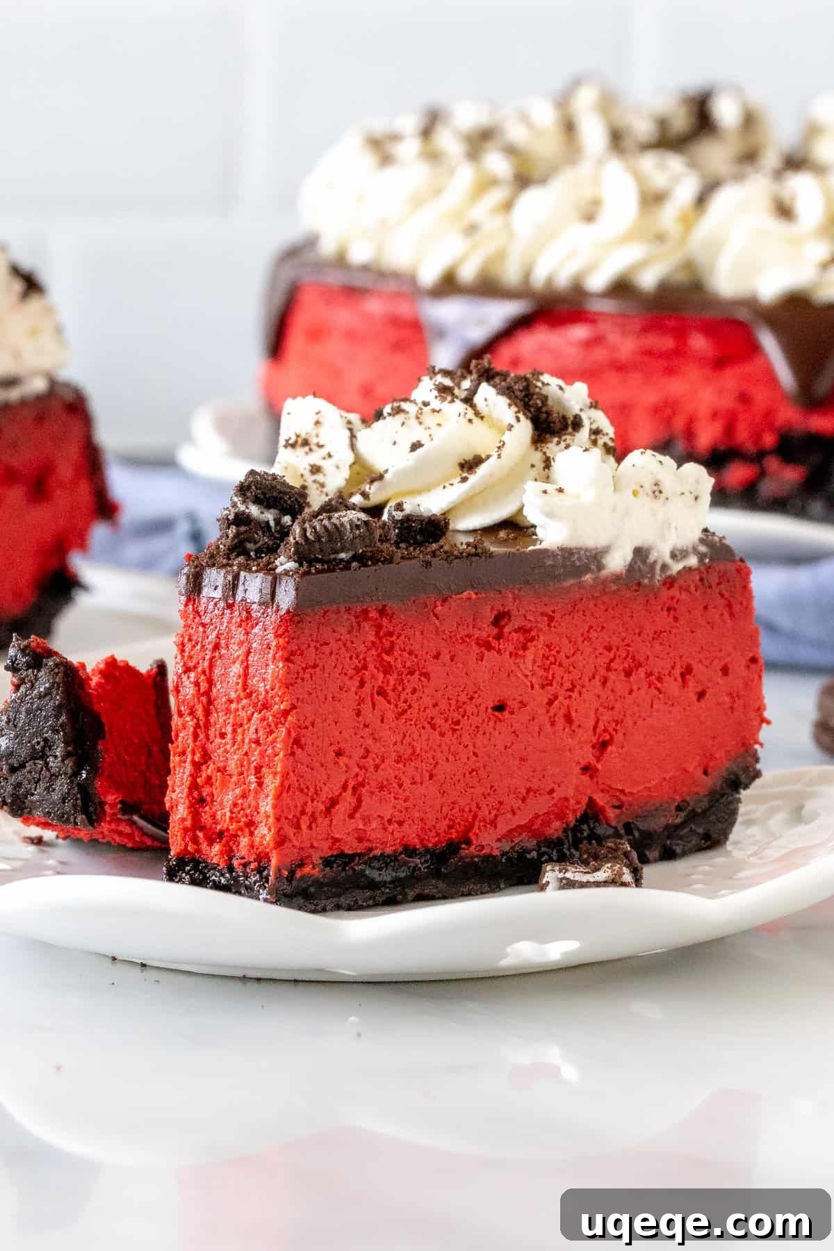 Velvety Oreo Dream Cheesecake 5 A close-up of a slice of Red Velvet Oreo Cheesecake with a small bite taken out, emphasizing the perfect creamy texture of the filling, the distinct Oreo crust, and the generous toppings of ganache, whipped cream, and crushed Oreos.