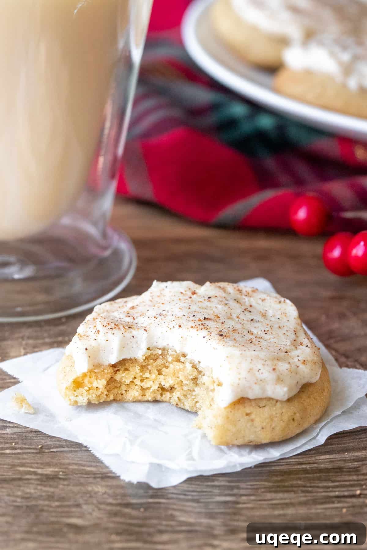 Spiced Eggnog Cookies with Creamy Frosting 3 A single frosted eggnog cookie with a bite taken out, resting beside a festive glass of eggnog, showcasing its soft interior.