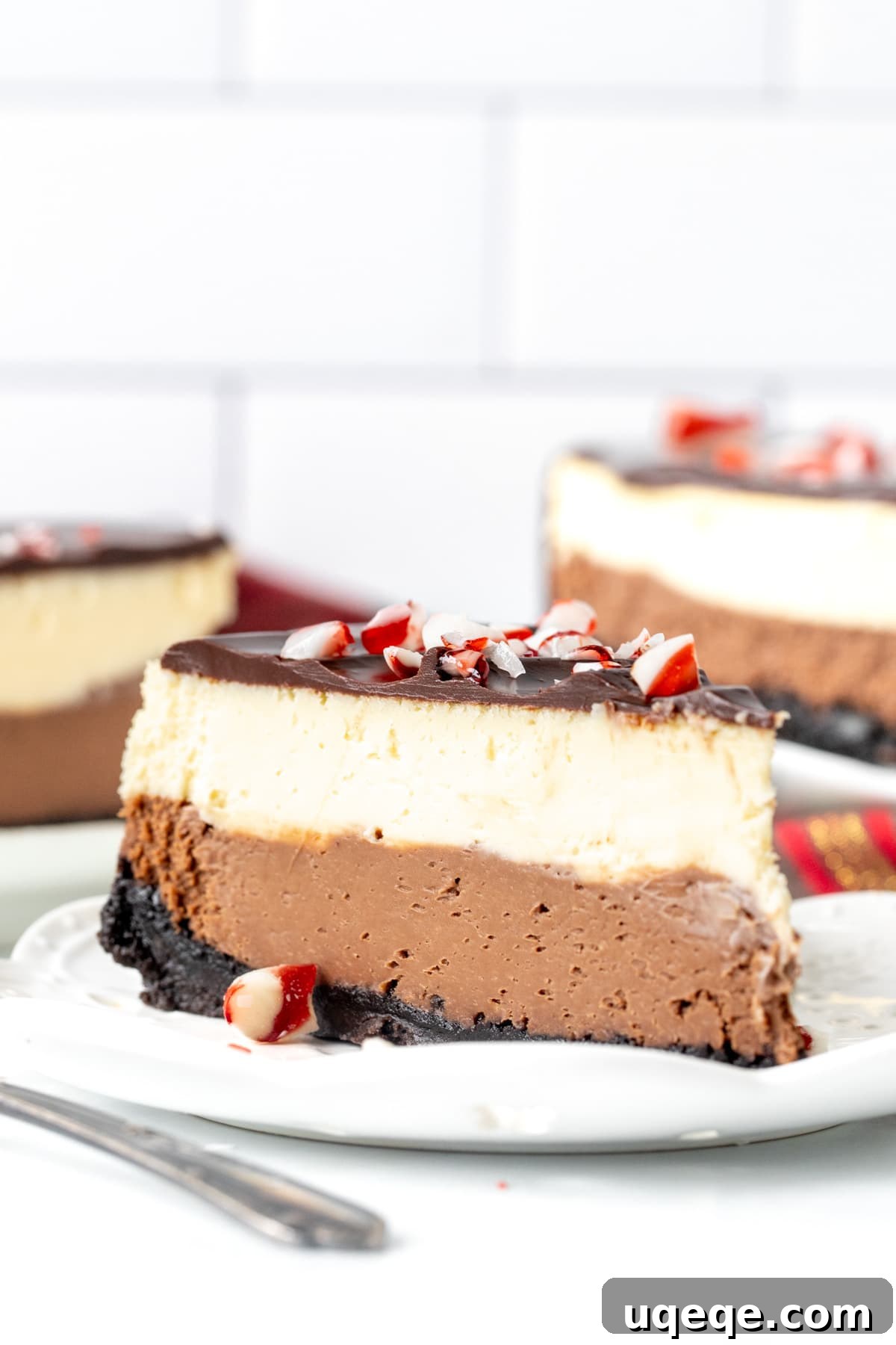 Candy Cane Crunch Cheesecake 3 A close-up view of a layered peppermint bark cheesecake slice, beautifully garnished with a smooth chocolate topping and sparkling crushed candy canes, presented on a white plate.