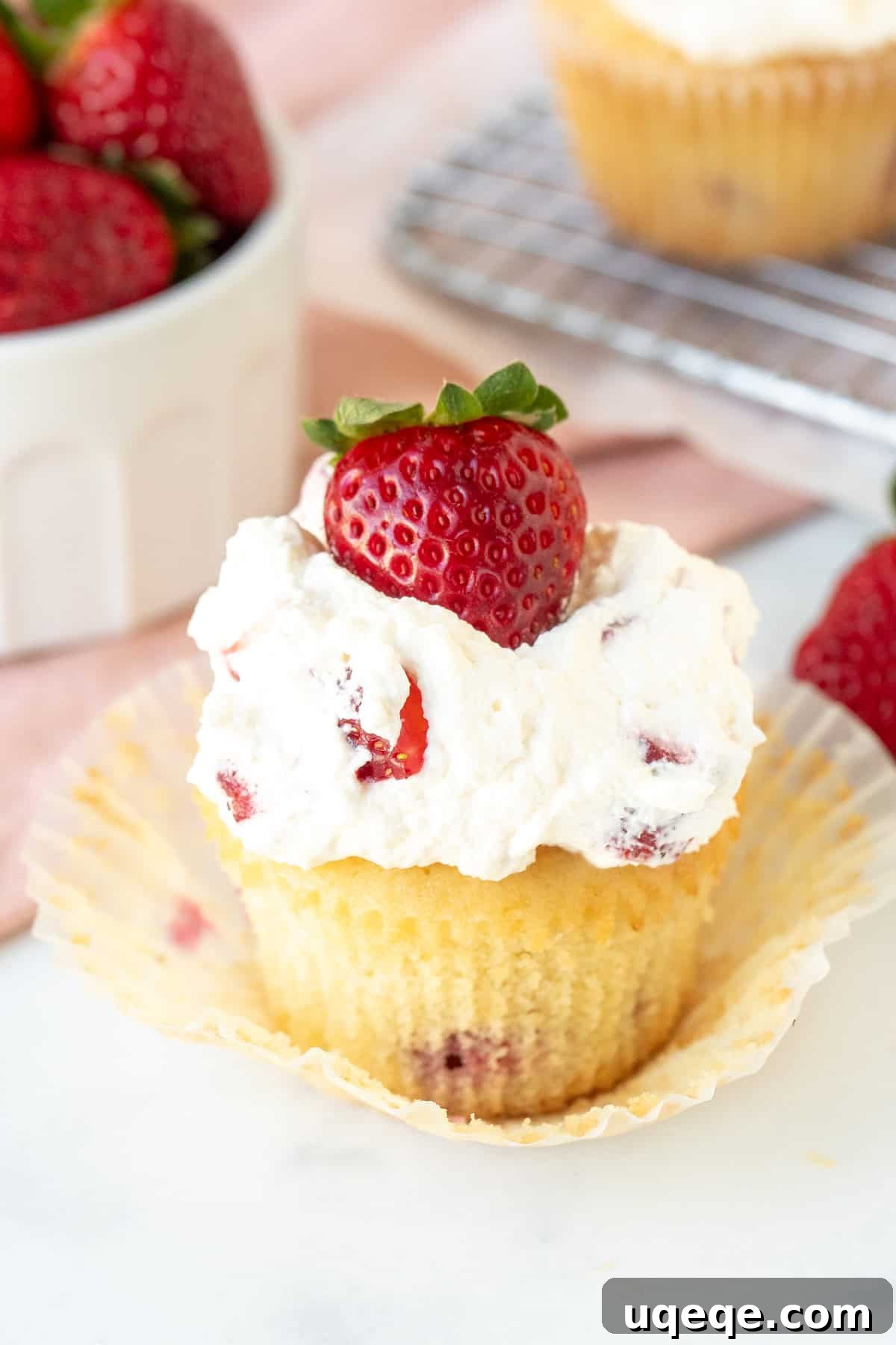 Luscious Strawberry Cream Cupcakes 2 A beautifully presented strawberries and cream cupcake with its muffin paper removed, set against a blurred background with a bowl of fresh strawberries.