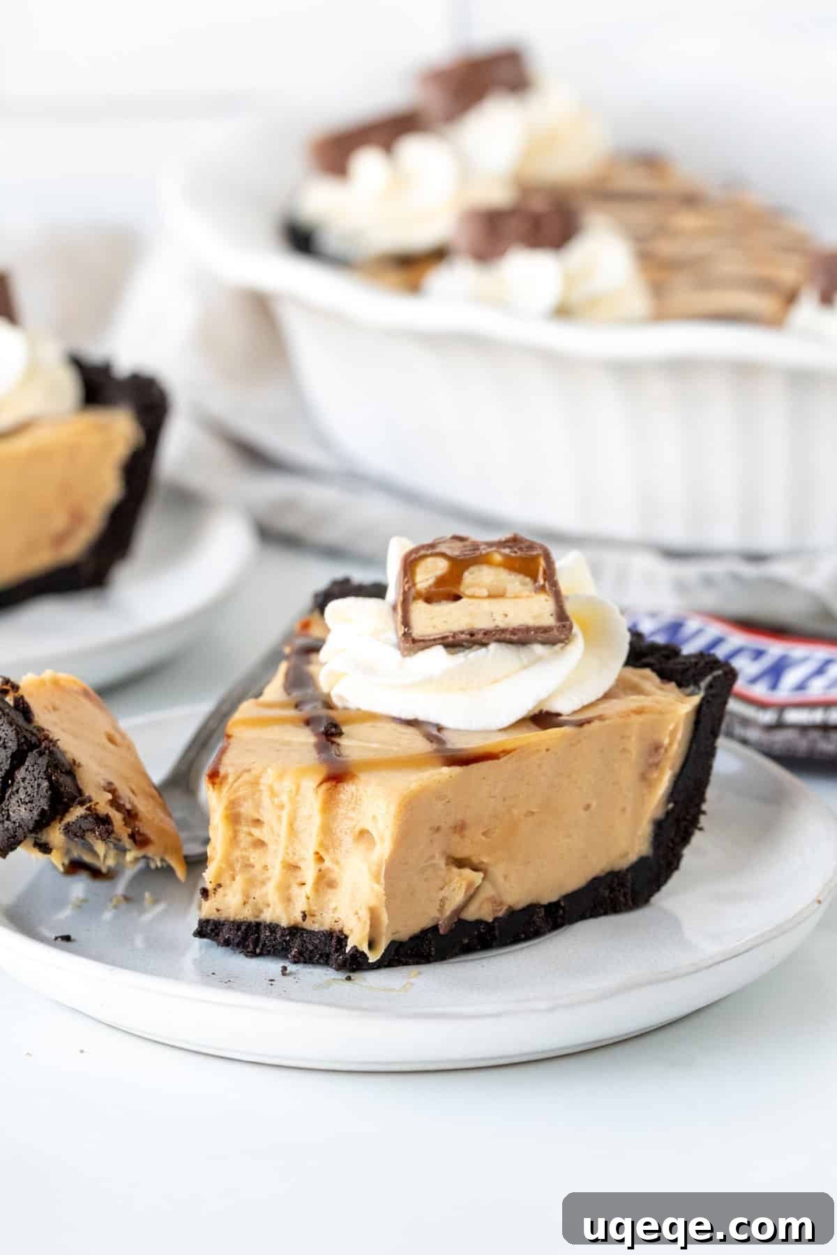 Decadent Snickers Pie 3 A close-up of a slice of Snickers pie, revealing its layered perfection: a dark Oreo crust, a thick, light brown peanut butter caramel filling dotted with Snickers pieces, and a generous topping of sauces.