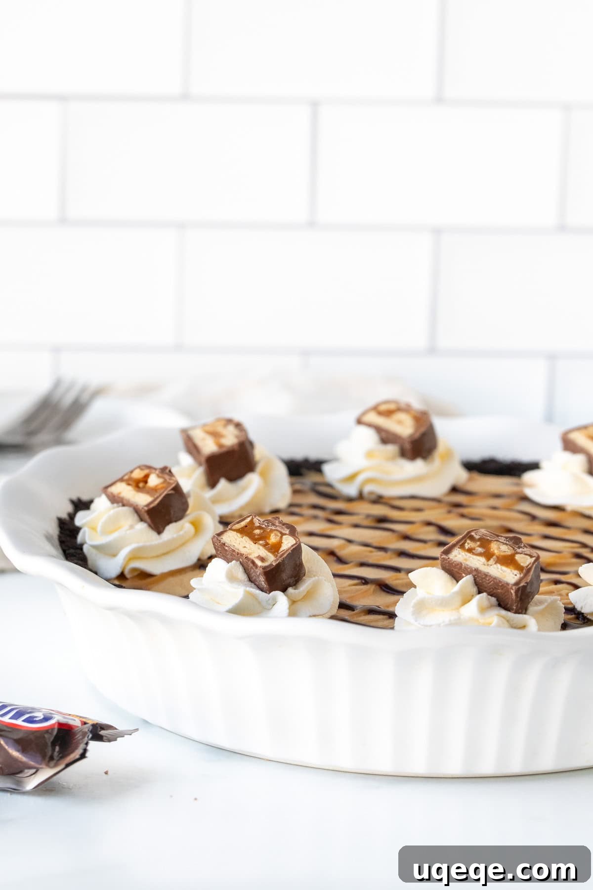 Decadent Snickers Pie 8 A beautifully garnished No-Bake Snickers bar pie, presented in a crisp white pie plate, topped with generous swirls of whipped cream, a rich caramel drizzle, and a scattering of finely chopped Snickers bars.
