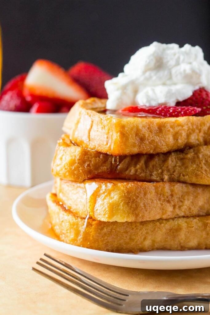 A tall stack of fluffy French Toast topped with whipped cream and strawberries, then drizzled in maple syrup.