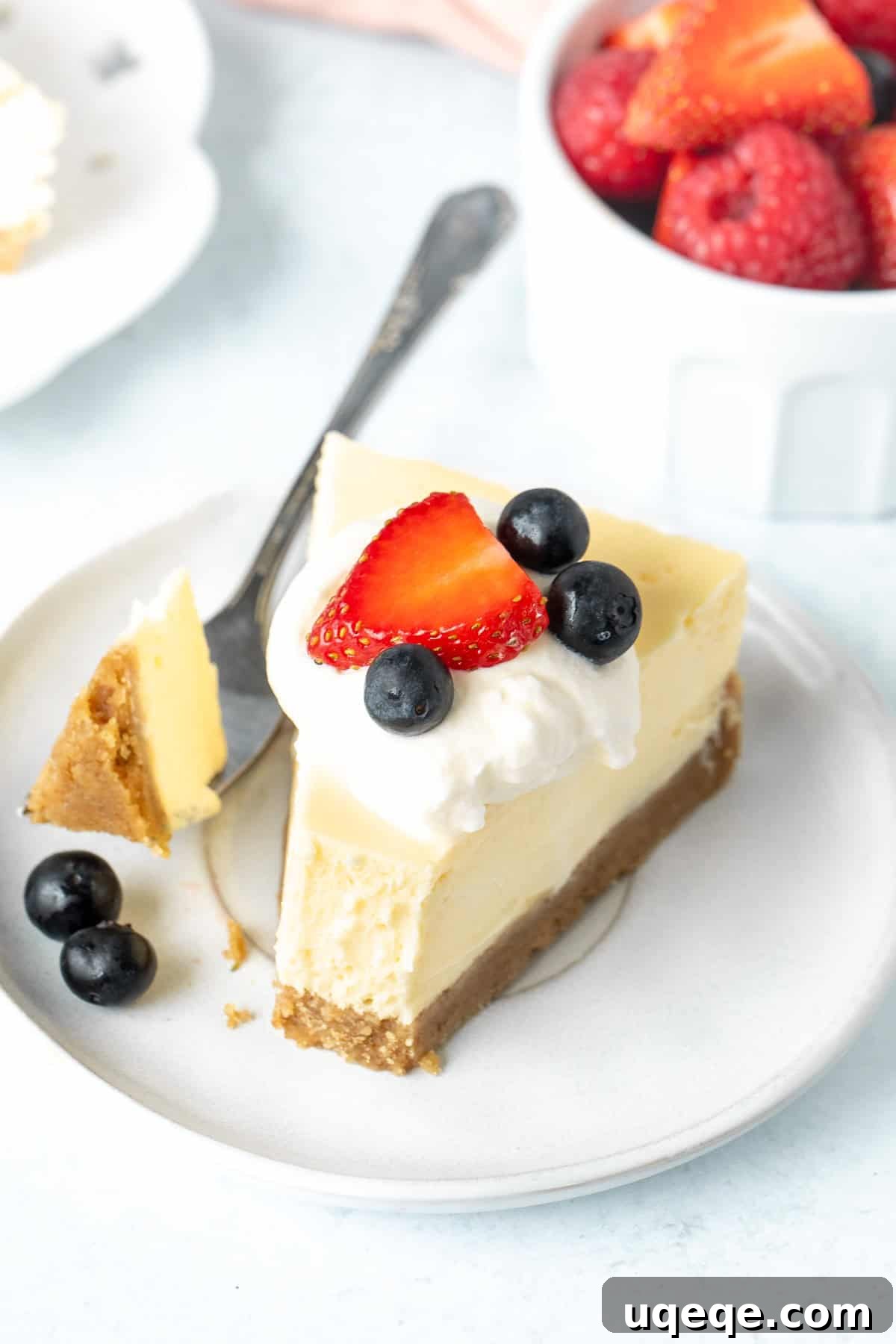 Intimate Batch Cheesecake 8 A perfectly sliced piece of small batch cheesecake topped with fresh whipped cream and berries, with a bite taken out.