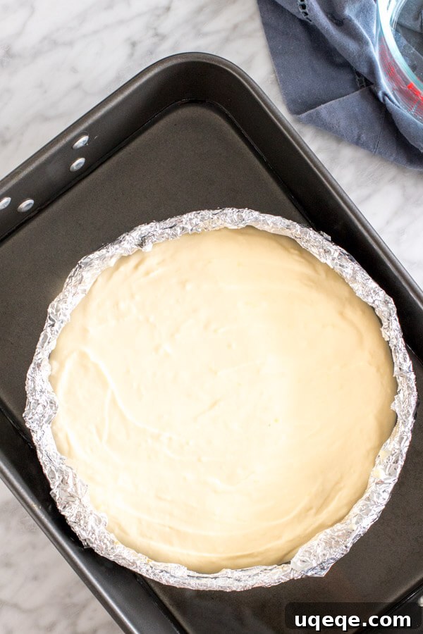 An unbaked lemon cheesecake mixture in a foil-wrapped springform pan, placed inside a large roasting pan filled with hot water for a water bath before baking.