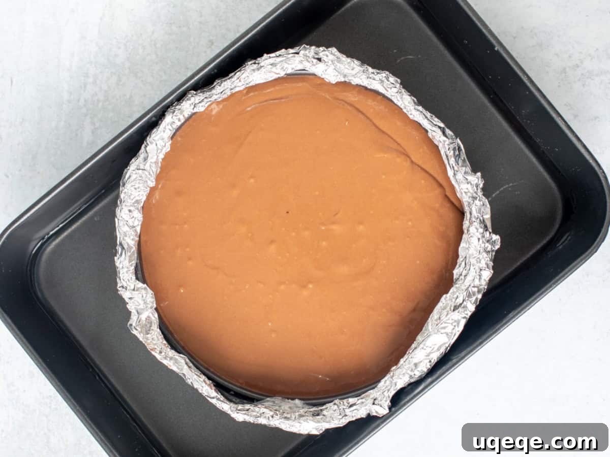 Cheesecake Water Bath Setup A springform pan containing cheesecake batter, wrapped in foil and placed inside a larger roasting pan filled with water, ready for baking.