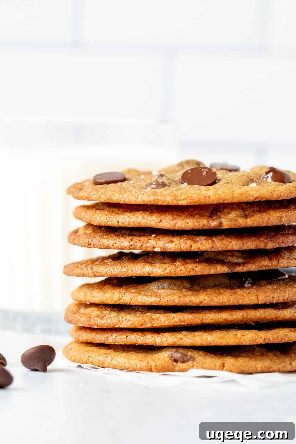 Stack of thin and crispy chocolate chip cookies with a glass of milk, highlighting their perfect golden edges and inviting texture