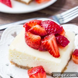Cheesecake bar topped with chopped strawberries on a white plate. Fork and second cheesecake bar in the background.