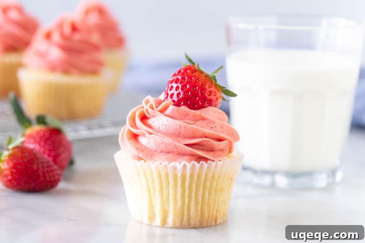 A beautifully decorated vanilla cupcake with generous swirl of fresh strawberry frosting, garnished with a sliced strawberry on top.