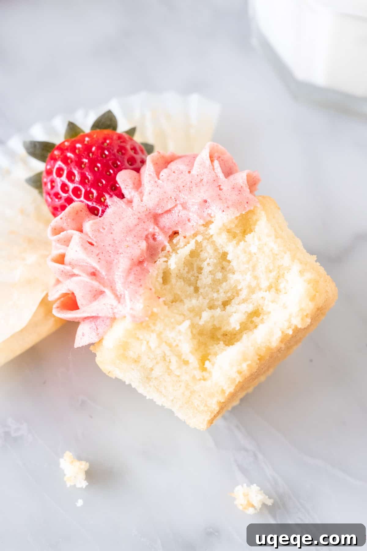 A vanilla cupcake with strawberry frosting, lying on its side, showing a bite taken out to reveal the moist cake and creamy interior.