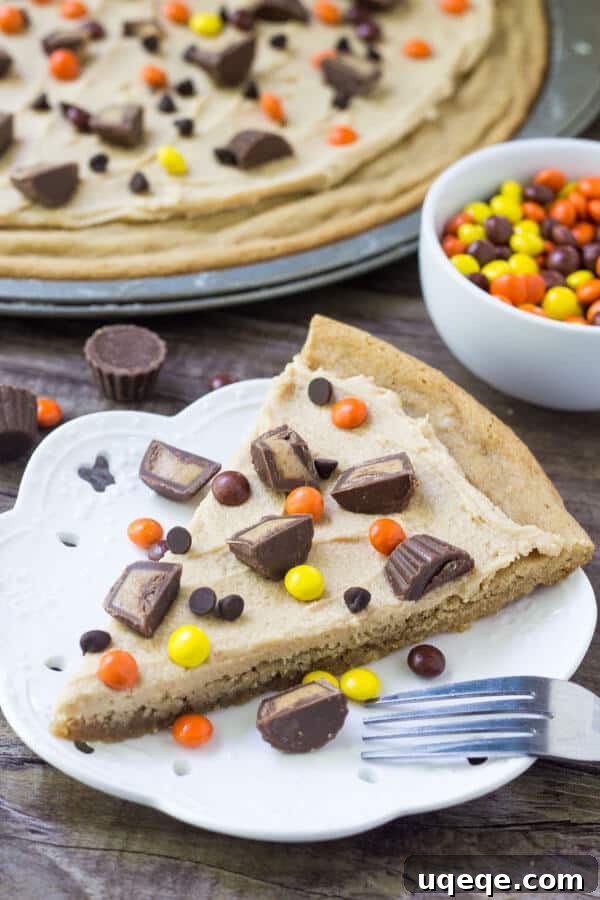 Slice of peanut butter cookie pizza topped with peanut butter frosting, peanut butter cups and peanut butter candies. Bowl of Reese's Pieces and whole cookie pizza in the background.