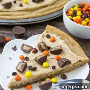 Slice of peanut butter cookie pizza topped with peanut butter frosting, peanut butter cups and peanut butter candies. Bowl of Reese's Pieces and whole cookie pizza in the background.