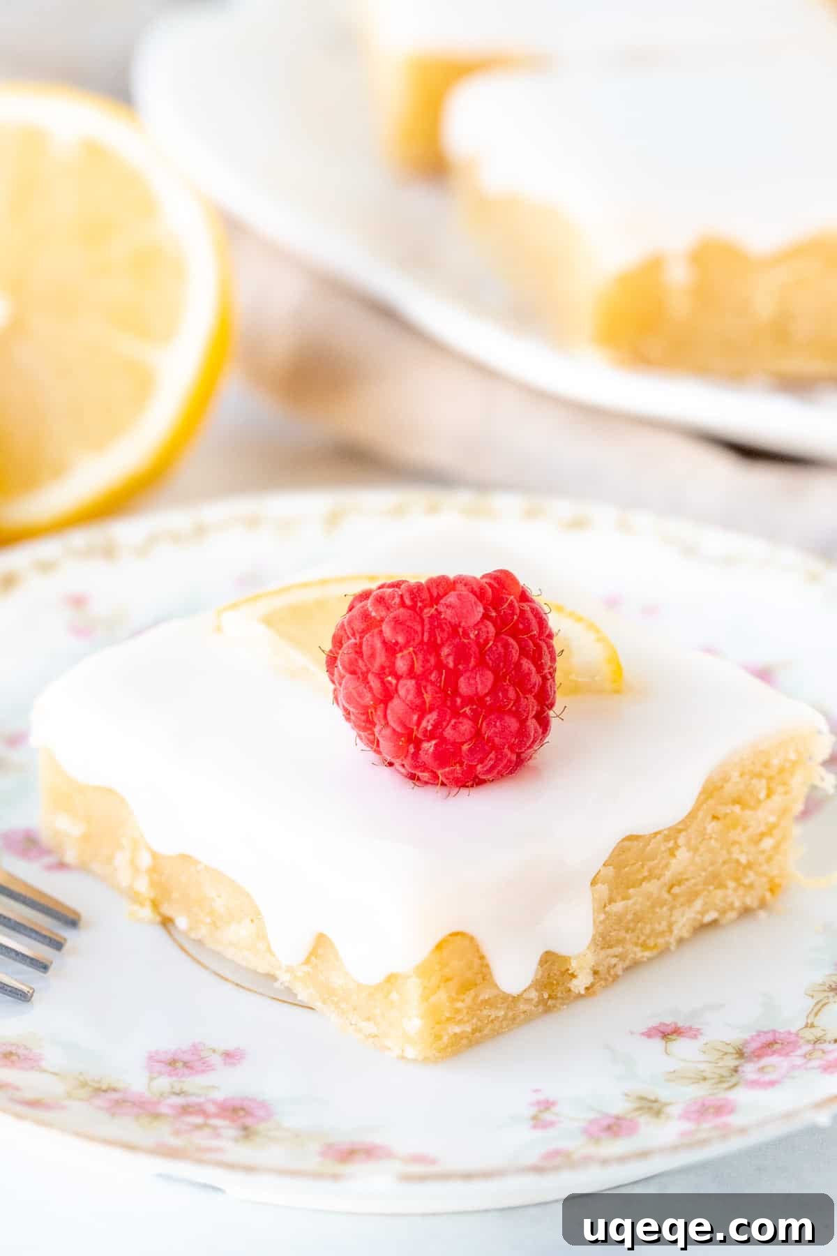 Zesty Lemon Blondies 7 A single glazed lemon brownie on a white plate, topped with a fresh raspberry and a thin lemon slice