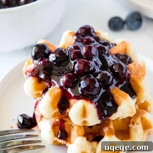 A stack of 2 small waffles topped with blueberry syrup with a bowl of blueberry sauce in the background.