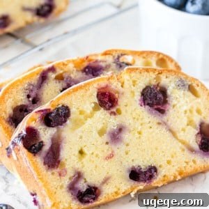 Three slices of moist blueberry bread on a cutting board, with the remaining loaf on a cooling rack in the background.