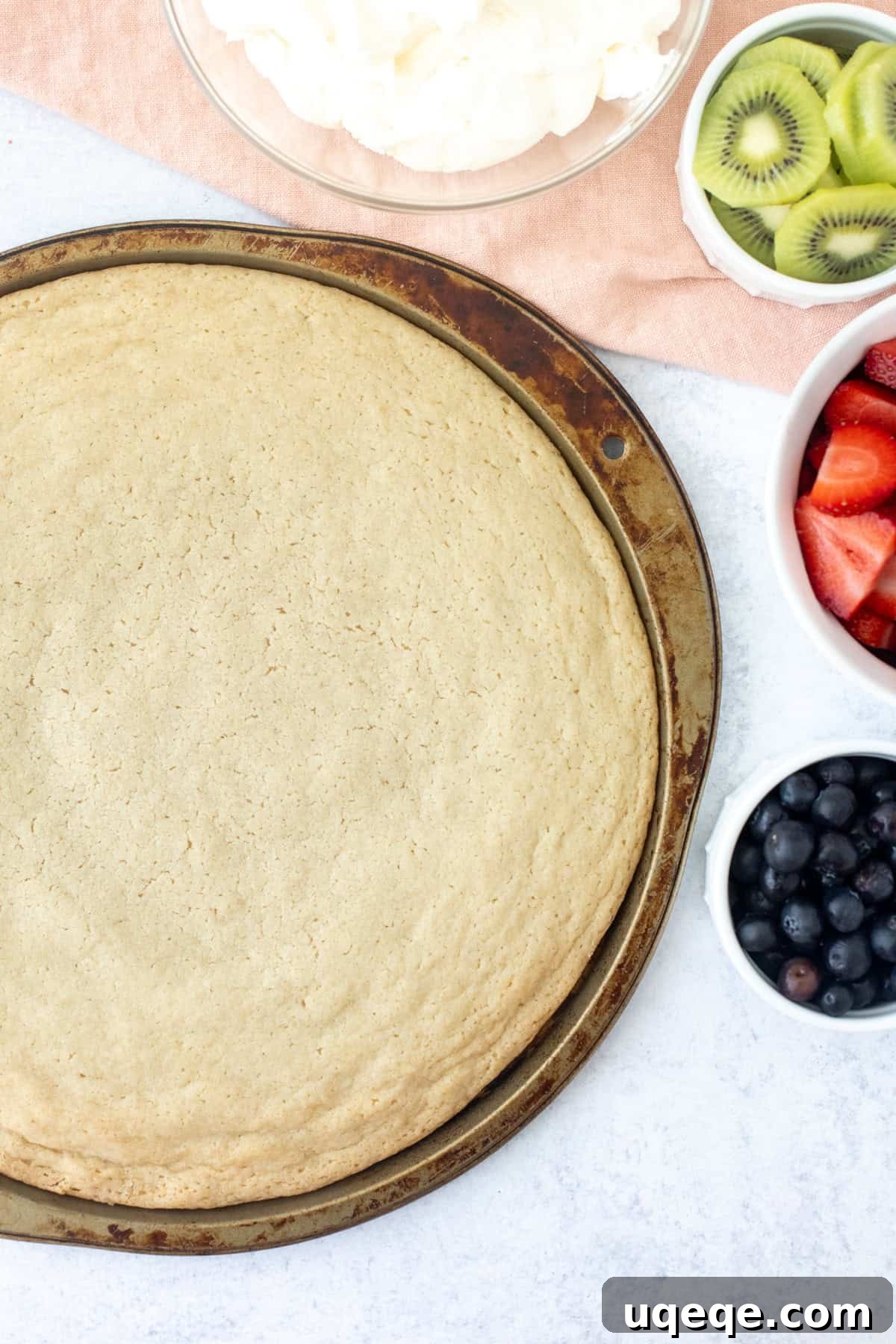 No-Bake Fruit Pizza with Cream Cheese Frosting 4 Sugar cookie made in a large pizza pan with bowl of whipped cream cheese frosting and bowls of fruit